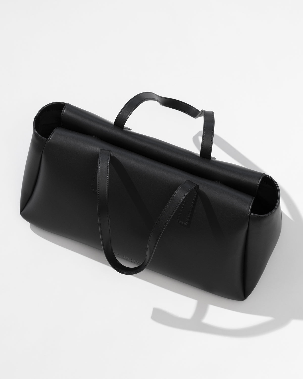 A black leather duffel bag with a structured, rounded rectangular shape, featuring dual top handles and a matching shoulder strap, photographed on a clean white background with a soft shadow.