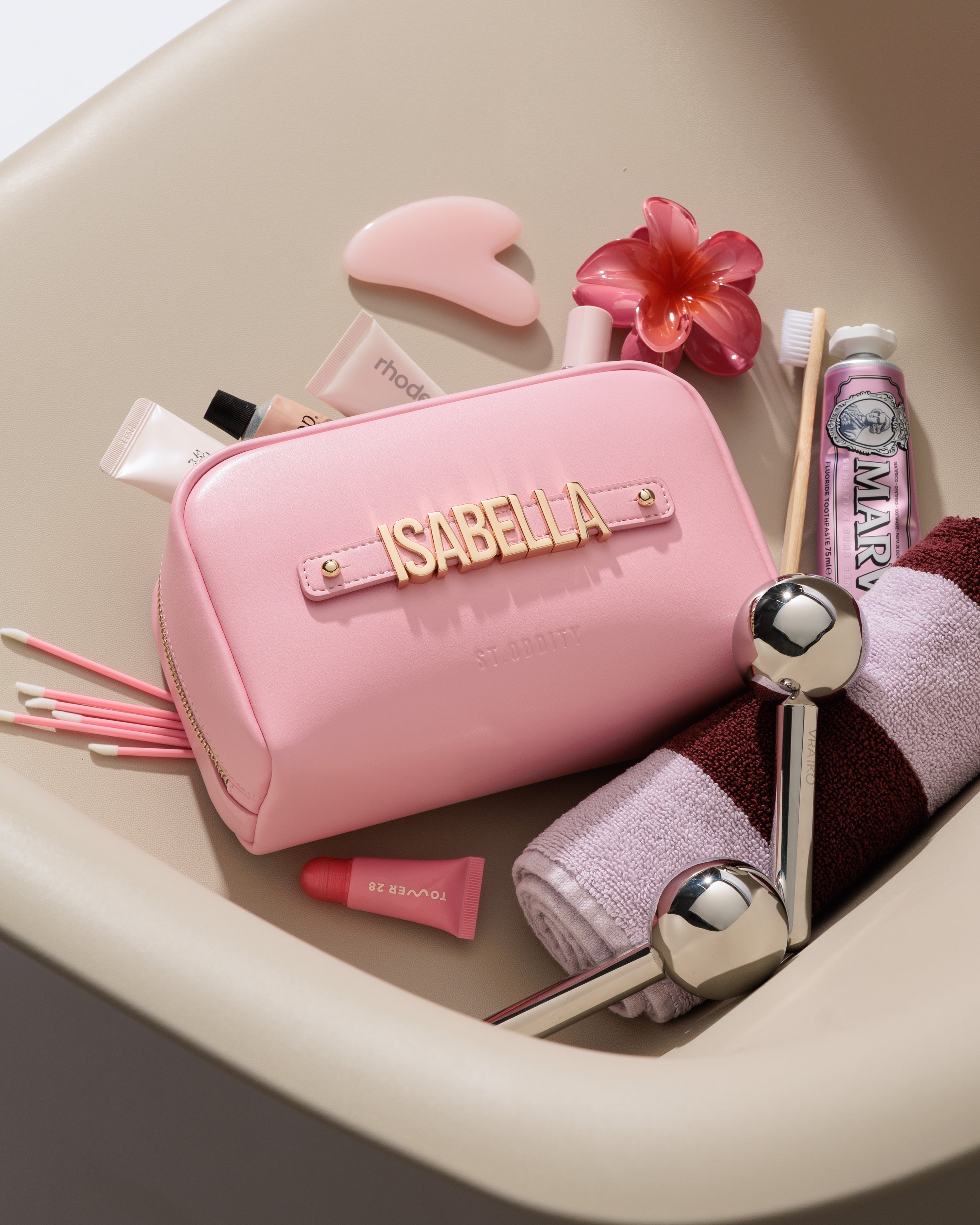Pink cosmetic pouch with 'ISABELLA' personalisation, surrounded by various beauty products on a beige surface.