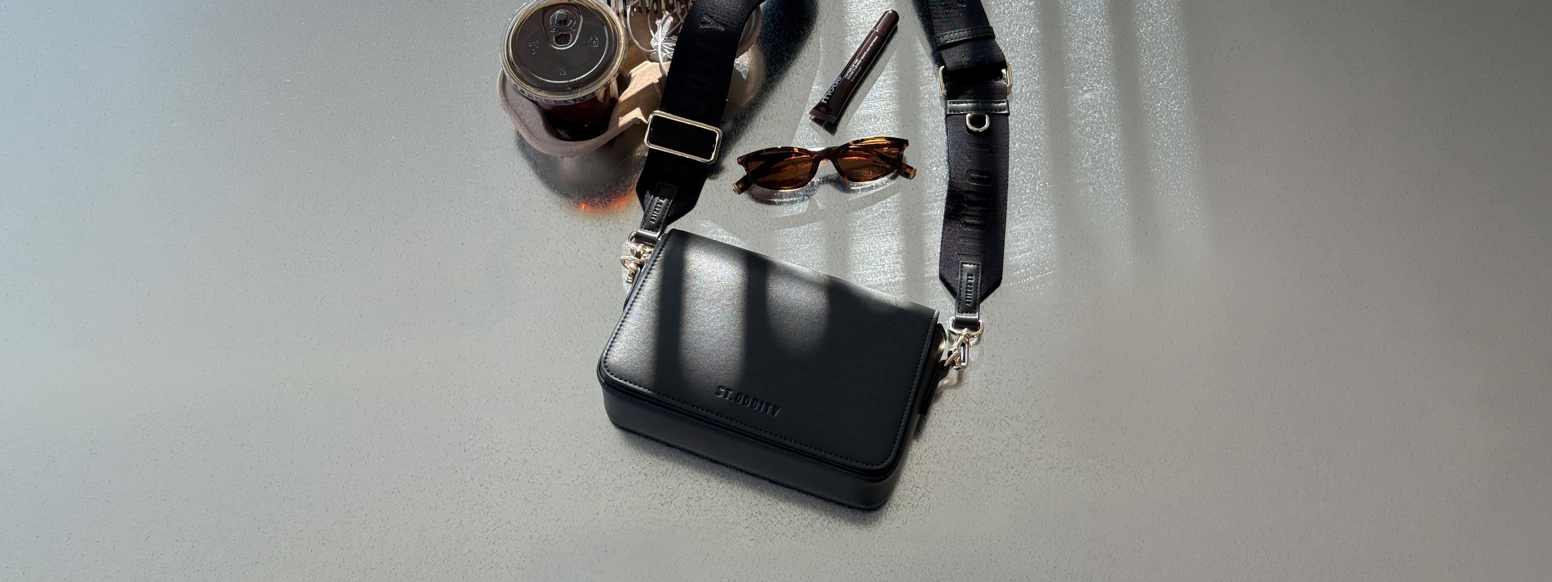 A black leather crossbody camera bag with a matching black strap, styled on a light gray surface with sunglasses, a pen, and a small bottle nearby.